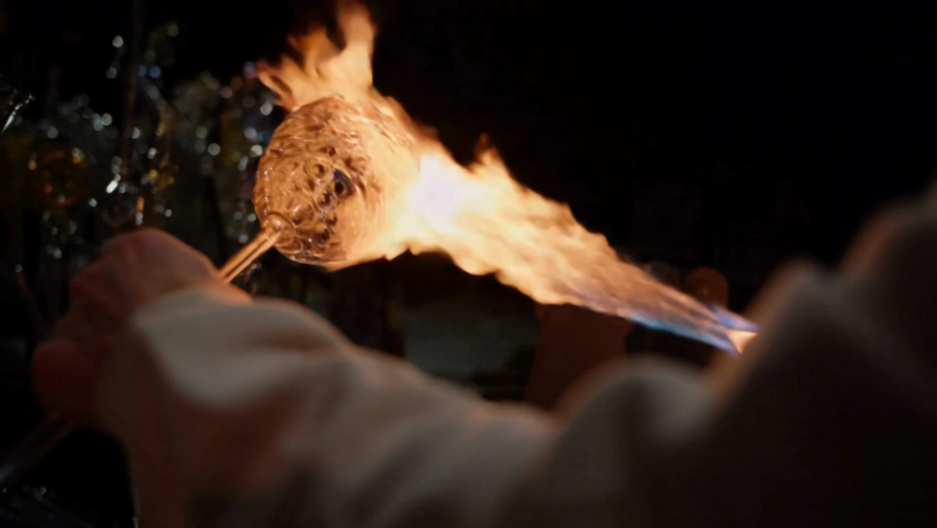The camera approaches the craftsman shaping glass by the fire